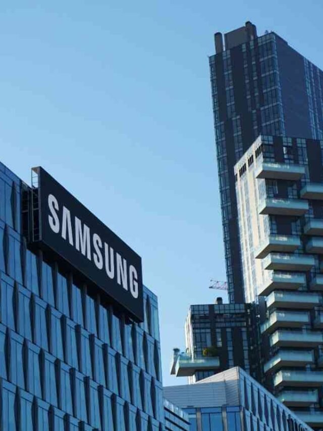 Photo by Capitalist Money on June 05, 2024. May be an image of The Samsung logo is displayed prominently on the side of a modern glass office building. The building has a unique design with angled glass panels. In the background, a taller, residential building with balconies is visible. The sky is clear and blue. The image may contain text such as 'SAMSUNG'.