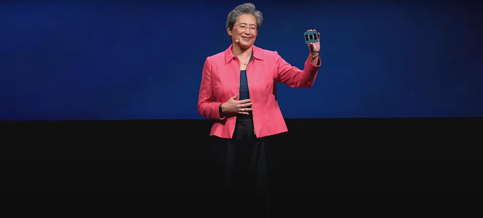 Photo by Capitalist Money on June 03, 2024. May be an image of A woman in a bright pink jacket stands on a stage with a blue backdrop, smiling at the audience while holding up a small, square object. .